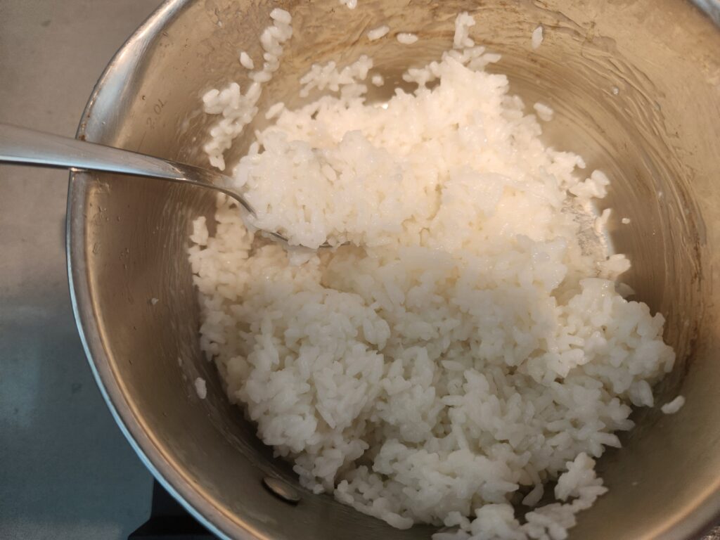 A picture of white rice in a stainless-steel pot with spoon. The rice is cooked well. It is light and fluffy in texture. 