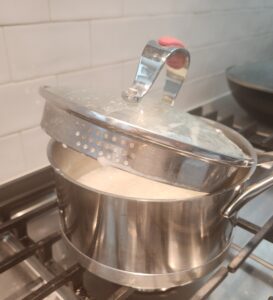 A stainless steel pot is on the stove with bubbling water and rice. The lid is slightly propped open.